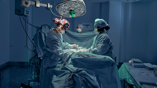 Navy surgeons in a brightly lit operating room, one wearing a patterned cap, performing surgery with a third surgeon observing in the background. 