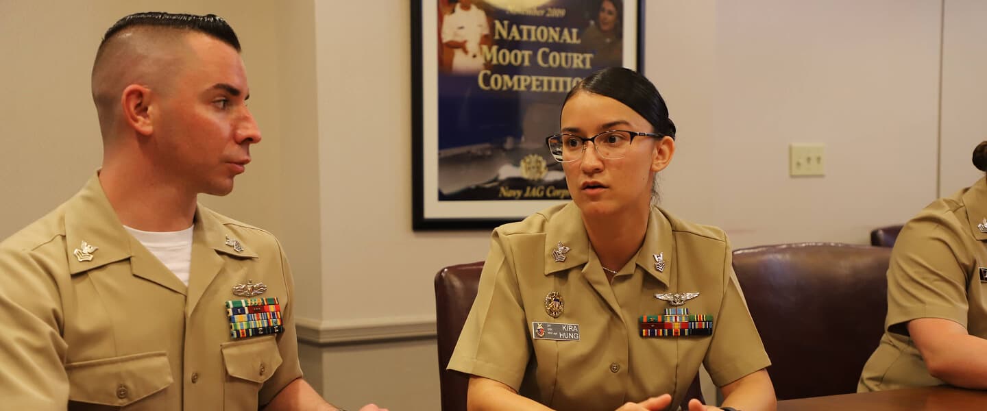 Navy Legalmen in khaki uniforms discuss legal matters at a table with an open book, a 'National Moot Court Competition' poster behind them