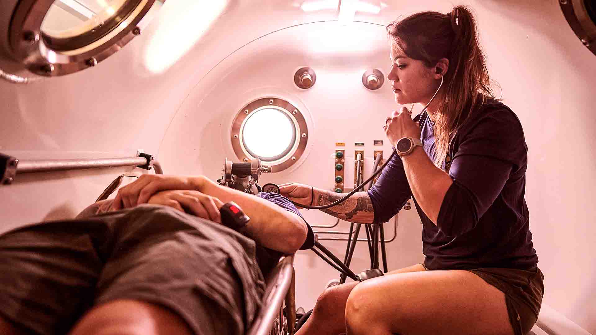 Navy Lieutenant Commander Mia Edgar in a hyperbolic chamber with a patient. She listens to his breathing with a stethoscope.